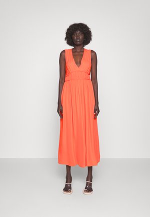 Maxi-mekko kirkkaan oranssina, jossa on syvä V-pääntie, kootut vyötärö ja virtaava kangas. Tyylitelty läpinäkyvillä korkokengillä.