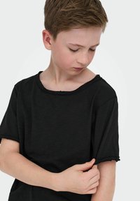 Czarny, krótki t-shirt z szerokim dekoltem i surowymi krawędziami, wykonany z teksturowanej tkaniny, trzymany w ręce blisko rękawa.