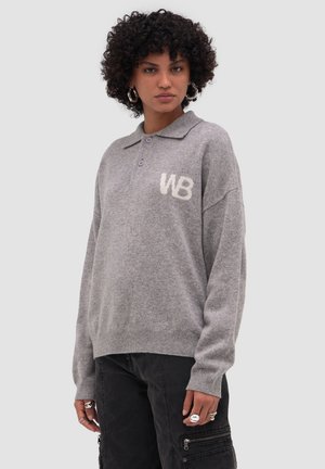 Kobieta o kręconych włosach, ubrana w szary sweter z dzianiny z kołnierzem i logo "WB", zestawiony z czarnymi spodniami cargo i srebrnymi kolczykami kołami.