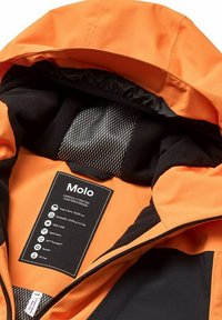 Oranje waterdichte jas met zwarte fleece voering, voorzien van een label met verzorgingsinstructies en technische specificaties. De getapete naden zijn zichtbaar.