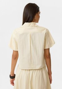 Korte mouwen, gekraagde shirt van zachte, lichte gele stof. Heeft een achterplooi, een gladde textuur en een losse pasvorm.