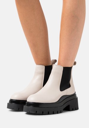 Bottines Chelsea en cuir beige avec un côté élastique noir, semelle épaisse noire et boucle décorative à l'arrière. Texture lisse et bout arrondi.