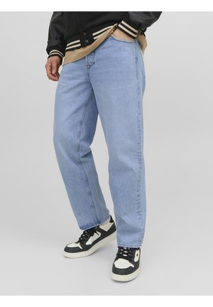 Persona con jeans rectos de color azul claro, zapatillas negras y blancas, una camisa beige y una chaqueta de universidad negra con puños a rayas.