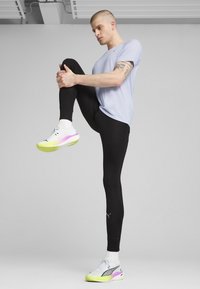 Sorte sportsleggings, lyserød t-shirt og hvide sneakers med gule og pink accenter. Modellen udfører en øvelse med hævet knæ.