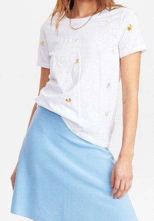 Femme portant un t-shirt blanc à manches courtes avec de petits papillons colorés brodés et une jupe bleu clair, debout avec la main posée.