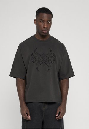 Mand står mod en ensfarvet baggrund iført en løs mørk T-shirt med et stort tribal-lignende design på brystet og mørke jeans.