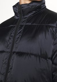 Chaqueta acolchada azul marino hecha de material brillante y acolchado. Cuenta con un cuello alto y una cremallera central con tirador negro.