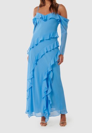 Kobieta ubrana w długą, jasnoniebieską sukienkę z odkrytymi ramionami, z falbanami i jednym przezroczystym długim rękawem, w połączeniu z beżowymi sandałami na obcasie.