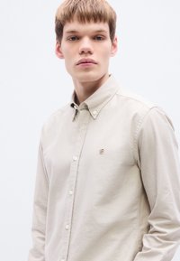 Εκρού μακρυμάνικο πουκάμισο με γιακά που κλείνει με κουμπιά, διακοσμημένο με μικρό κεντημένο λογότυπο στο στήθος και τυπική εμπρόσθια σειρά κουμπιών.