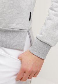 Sudadera gris con una textura suave, con una abertura lateral y puños acanalados. Destacada con una pequeña etiqueta negra en el dobladillo.