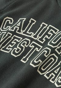 Tamnozelena tkanina majice s velikim bijelim natpisom "CALIFORNIA WESTCOAST." Tekstura je glatka s blagim rastezanjem.