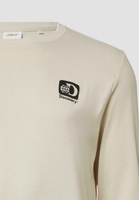 Beżowa koszula z długim rękawem, wykonana z miękkiego materiału, z czarnym logo "Discovery" z obrazkiem globu na górnej lewej części klatki piersiowej.