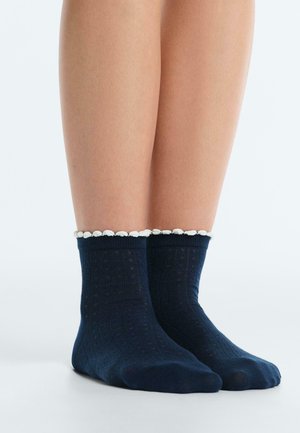 Nohy v tmavomodrých členkových ponožkách so subtílnym textúrovaným vzorom a bielou zúbkovanou lemovkou hore, na jednotnom pozadí.