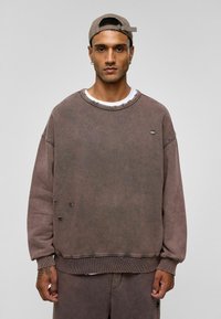 Overmaatse bruine sweatshirt met een ronde hals, versleten details en geribbelde manchetten. Gecombineerd met een bijpassende onderkant, met rafelige randen.