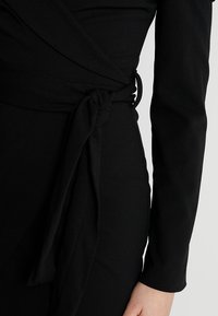Black wrap dress featuring long sleeves, a tied waist detail, and a smooth, textured fabric. The fabric appears stretchy and fitted.