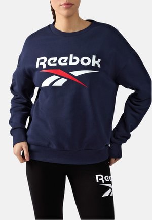Vrouw draagt een marineblauwe Reebok-trui en zwarte legging met het Reebok-logo op de linker dij, tegen een effen achtergrond.