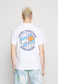Hvid bomulds t-shirt med et flerfarvet fredstegn grafisk motiv på bagsiden, med teksten "KOM SAMMEN VERDEN OVER" i marineblå.
