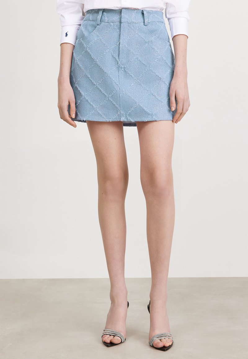 Femme portant une mini-jupe texturée bleu clair avec des poches latérales, associée à une chemise blanche et des talons ouverts sur un fond uni.