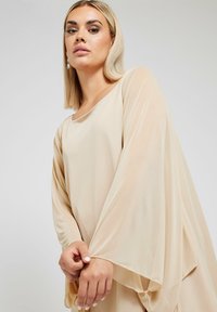 Ljusbeige klänning med långa, flödande ärmar och en lös silhuett. Tyget ser mjukt ut med en slät textur och en mjuk drapering.
