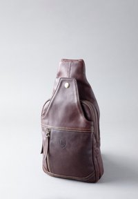Brown leather sling bag with a smooth texture, featuring a rounded shape, side zip pocket, and a brass snap detail on the top.