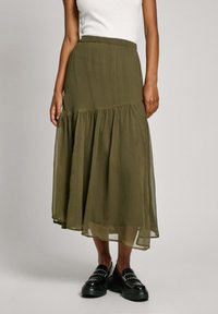 Jupe midi à volants en vert olive, fabriquée dans un tissu léger et transparent. Taille élastique avec une silhouette fluide et un ourlet inférieur superposé. Associée à des chaussures noires.
