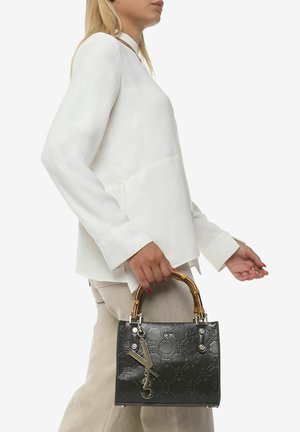 Zwarte gepatineerde leren handtas met reliëflogo-patroon, bamboe handgreep en zilveren hardware-accenten, vastgehouden door een persoon in een witte blouse.
