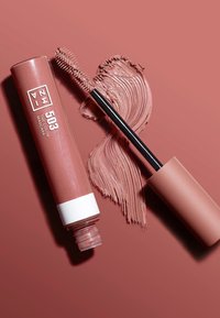 Färgad mascara i en cylindrisk tub; mjuk rosa, med en svart borste med borst; krämig produktprov bredvid.