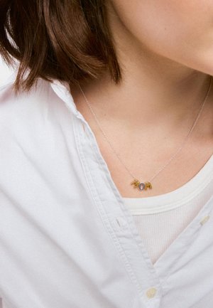 Mujer que lleva un delicado collar de cadena con letras de oro y plata que forman la palabra "MOM" sobre una camisa blanca y una camiseta sin mangas.