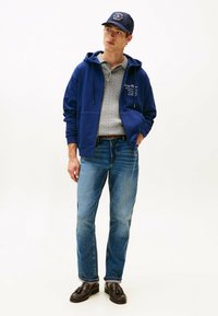 Felpa blu con zip sopra un polo in maglia grigio chiaro, abbinata a jeans azzurri e mocassini marroni. Il cappello presenta un logo. Look semplice e casual.