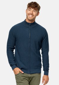 Sudadera con cremallera de color azul oscuro y cuello alto, con un patrón de punto texturizado, mangas largas y puños ajustados. Lleva puesta sobre pantalones de color verde oliva.