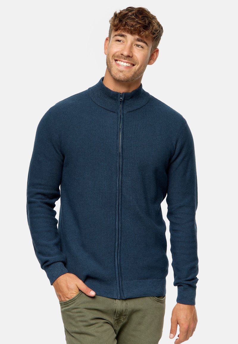 Sudadera con cremallera de color azul oscuro y cuello alto, con un patrón de punto texturizado, mangas largas y puños ajustados. Lleva puesta sobre pantalones de color verde oliva.