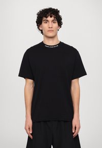 Melnas kokvilnas T-krekls ar apaļu kaklu un kreisajā krūtī kabatu. Kakla izgriezumam ir balts "VERSACE JEANS COUTURE" zīmols.