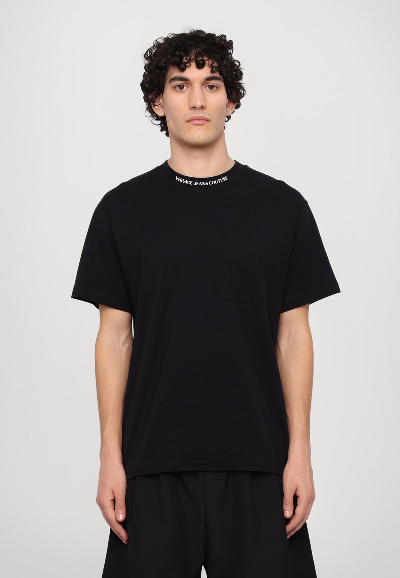 Melnas kokvilnas T-krekls ar apaļu kaklu un kreisajā krūtī kabatu. Kakla izgriezumam ir balts "VERSACE JEANS COUTURE" zīmols.