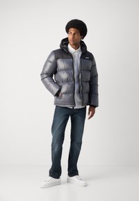Pufferjakke i gradient grå med hette, med glidelås foran, elastiske mansjetter og logo på venstre bryst; kombinert med blå jeans og hvite sneakers.