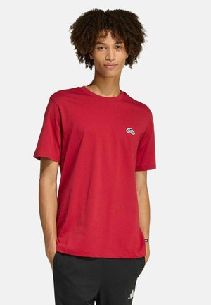 Jonge man met krullend haar, draagt een rood T-shirt met een klein logo en zwarte broek met een wit logo, staand tegen een effen achtergrond.