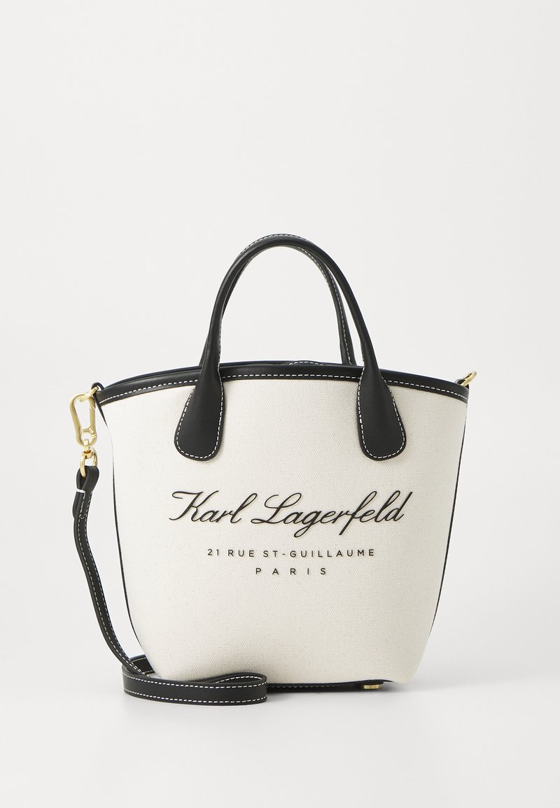Valge kangast käsitasku mustade nahkdetailidega, ülemised käepidemed ja reguleeritav õlarihm. Esiküljel on must prinditud brändi logo.