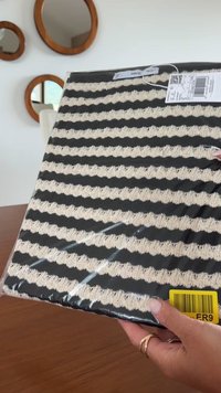 Mani che tengono un capo di abbigliamento piegato a righe bianche e nere avvolto in plastica con etichette su un tavolo di legno, con specchi tondi attaccati al muro dietro.