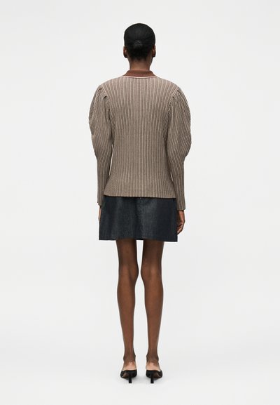 Άτομο με τα μαλλιά σε κότσο φοράει ένα καφέ ριμπ πλεκτό πουλόβερ με φουσκωτούς ώμους, μαύρη φούστα και μαύρα παπούτσια με τακούνια, στέκεται γυρισμένο πλάγια.