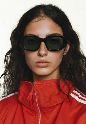 Femme aux cheveux bouclés foncés portant des lunettes de soleil noires rectangulaires et une veste rouge à fermeture éclair avec des rayures blanches sur les épaules.