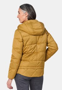 Puffjacke in senfgelb mit Kapuze, versehen mit einem gesteppten Design, glatter Textur und einem etwas längeren Rücken für zusätzlichen Schutz.