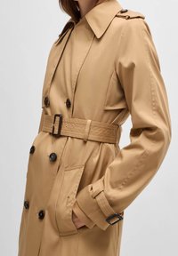 Kobieta w beżowym płaszczu trench z czarnymi guzikami, z ręką w kieszeni, na jednolitym jasnoszarym tle.