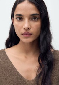 Maglione marrone a maglia con scollatura a V, texture morbida. Capelli lunghi, scuri e ondulati. Trucco naturale e neutro che mette in risalto occhi e labbra.