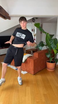 Junger Mann in schwarzen Shorts und grafischem T-Shirt hebt sein Shirt und zeigt den Bauch, steht auf einem Holzboden in einem hellen Wohnzimmer mit großer Pflanze und orangem Sofa.