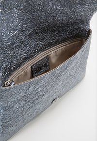 Metaliczna srebrna torebka o teksturowanej powierzchni, z wewnętrzną kieszonką na zamek błyskawiczny i metką z marką. Ma gładką, materiałową podszewkę.