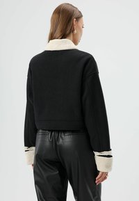 Zwarte cropped jas met crème faux shearling kraag en manchetten, gladde textuur en contrasterende stiksel details. Gecombineerd met zwarte broek.