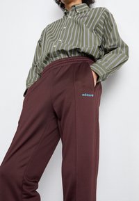 Vinröda Adidas-joggers med elastisk midjerem och blå logga. Kombinationen inkluderar en grön randig skjorta med vita vertikala ränder.