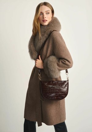 Kobieta ubrana w brązowy wełniany płaszcz z dużym futrzanym kołnierzem i mankietami, trzymająca ciemnobrązową torebkę na ramię ze skóry o fakturze krokodyla.