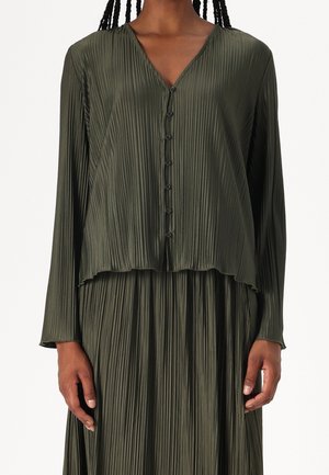 Donkergroene geplooide blouse met lange mouwen en knopen, gecombineerd met een bijpassende donkergroene geplooide rok, gedragen door een staand persoon.