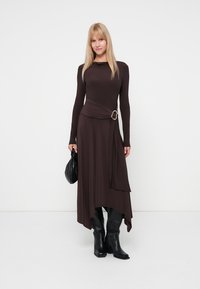Mulher vestida com um vestido castanho escuro de mangas compridas, com um hem assinalado e detalhe de fivela, segurando uma mala preta e usando botas pretas.