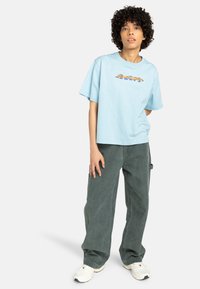 T-shirt en coton bleu clair avec un motif coloré sur le devant, associé à un pantalon large vert foncé et des baskets blanches.